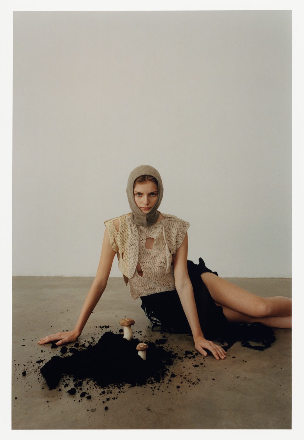 Woman in textured top and head covering sits on the floor beside pile of soil and mushrooms against a neutral wall backdrop.