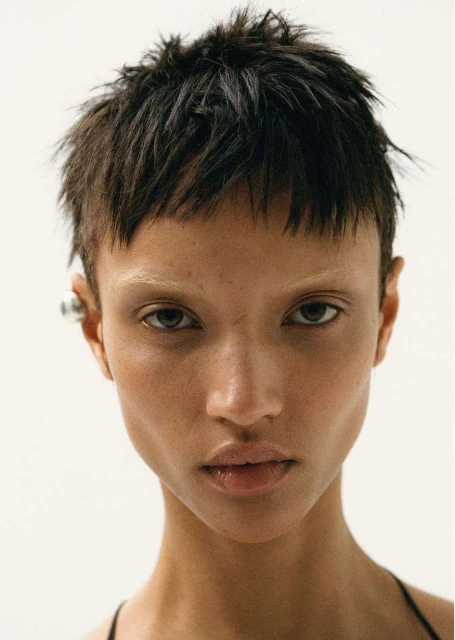 Close-up portrait of a woman with short, dark hair, light eyebrows, and a serious expression against a plain white backdrop.