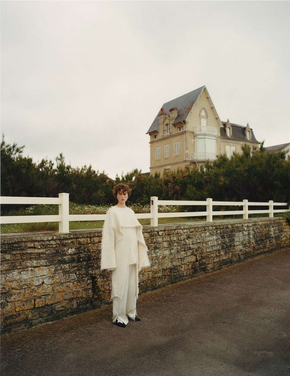 Adult woman in stylish cream-colored clothes standing on a road near a brick wall with a white fence and a large country house.