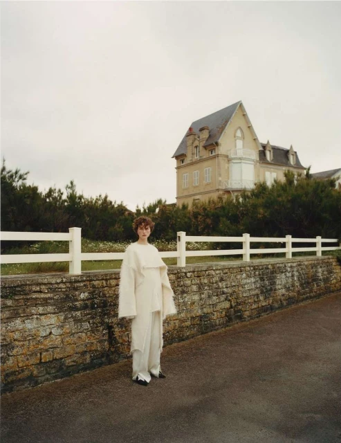 Adult woman in stylish cream-colored clothes standing on a road near a brick wall with a white fence and a large country house.