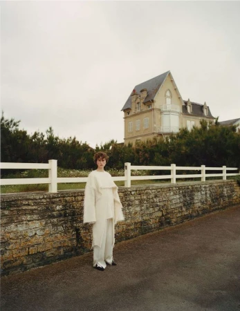 Adult woman in stylish cream-colored clothes standing on a road near a brick wall with a white fence and a large country house.