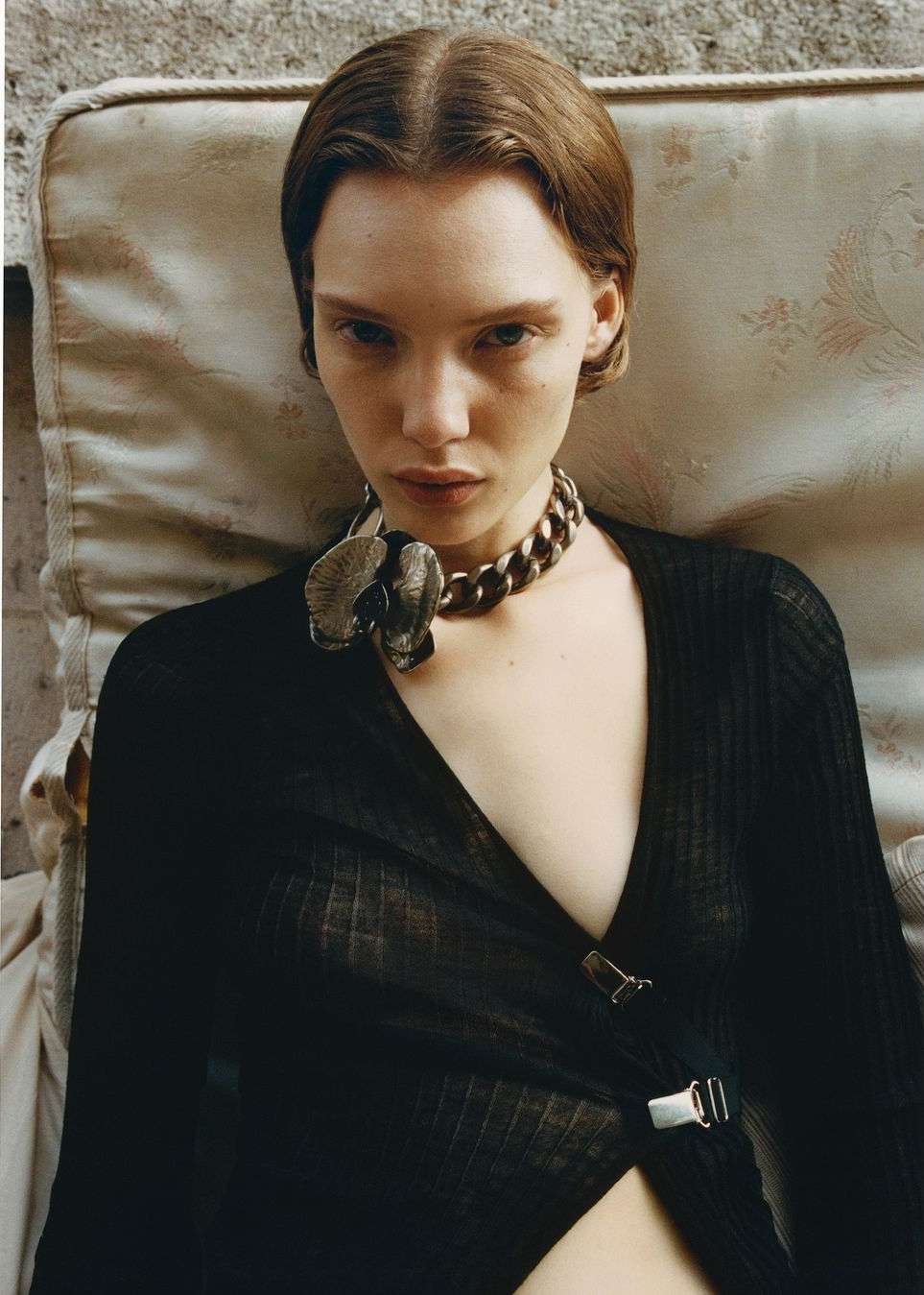 Close-up portrait of a young woman with brown hair and fair skin, wearing a black top and a chunky metal necklace with a flower pendant.