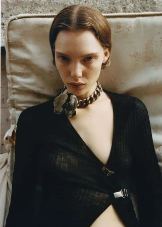 Close-up portrait of a young woman with brown hair and fair skin, wearing a black top and a chunky metal necklace with a flower pendant.