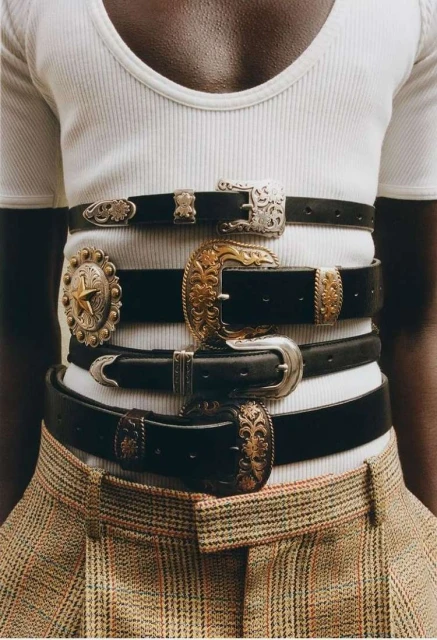 Upper torso shot of a person wearing a white shirt and multiple black leather belts with decorative metal buckles over plaid pants.