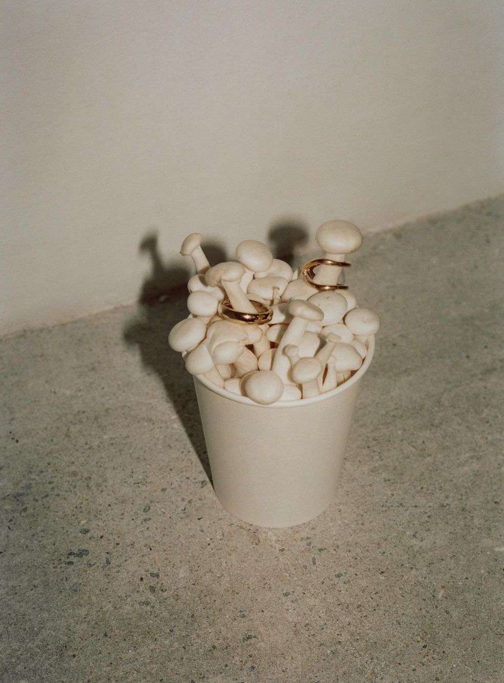 Still life of shimeji mushrooms in a paper cup, adorned with gold rings and ear cuffs, against a neutral wall and speckled floor.
