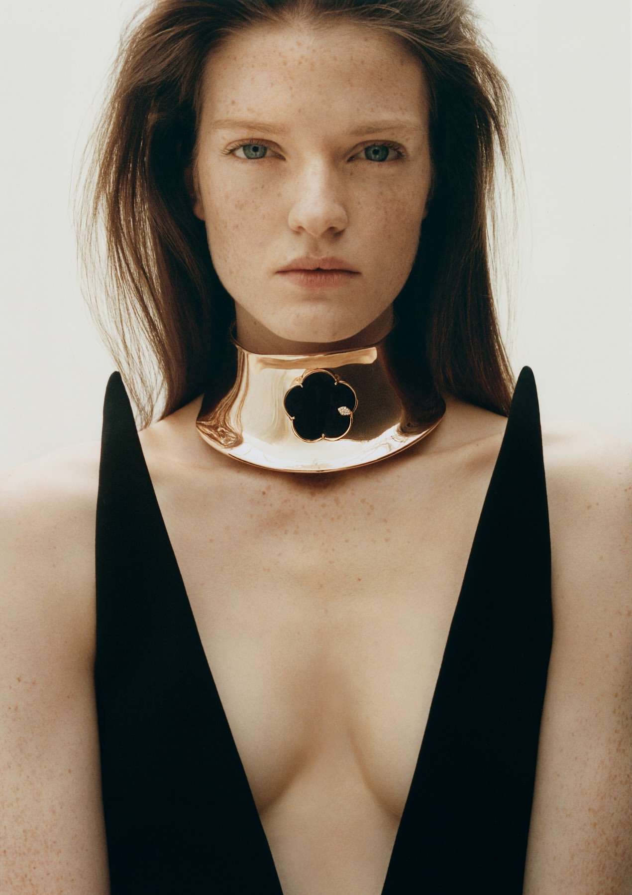Portrait of a woman with long brown hair and pale skin, wearing a black top and a gold choker necklace with a black flower detail.