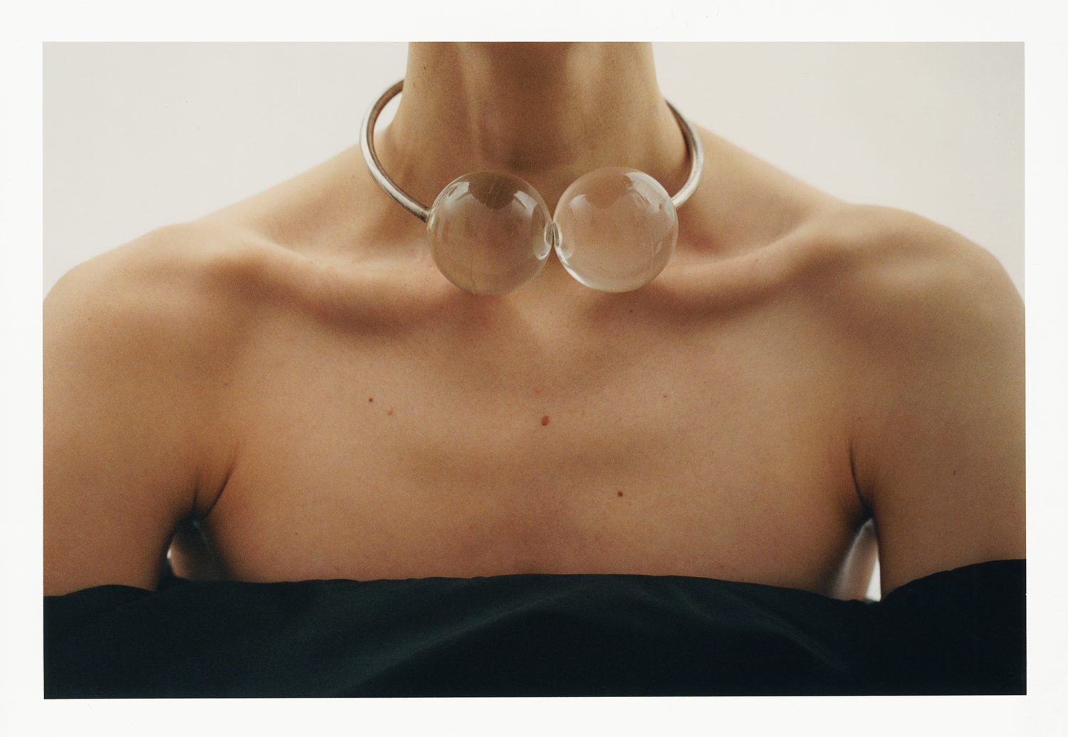 Close-up of a woman's shoulders and chest wearing a silver collar necklace with two clear glass spheres against a white backdrop.