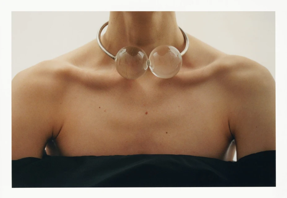 Close-up of a woman's shoulders and chest wearing a silver collar necklace with two clear glass spheres against a white backdrop.