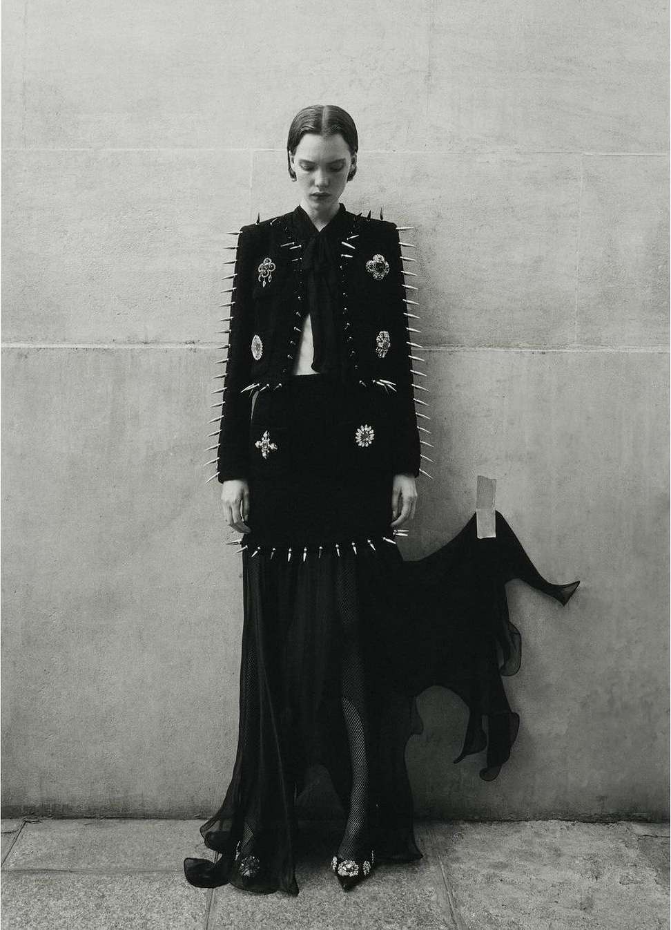 A lone fashion model poses against a neutral wall in a black jacket, skirt, and long dress accented with silver spikes and embellishments.