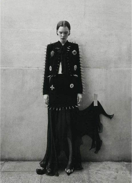 A lone fashion model poses against a neutral wall in a black jacket, skirt, and long dress accented with silver spikes and embellishments.