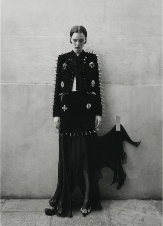 A lone fashion model poses against a neutral wall in a black jacket, skirt, and long dress accented with silver spikes and embellishments.