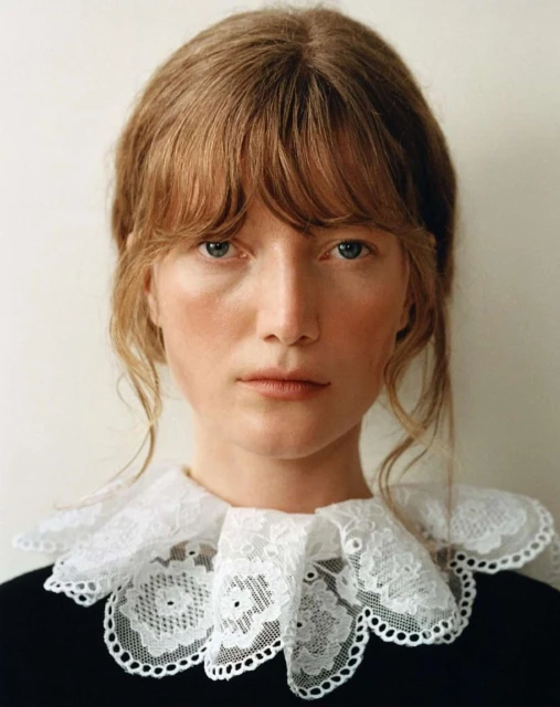 Close-up portrait of an adult female with light brown hair and blue eyes, wearing a black top and a white lace collar against a white background.