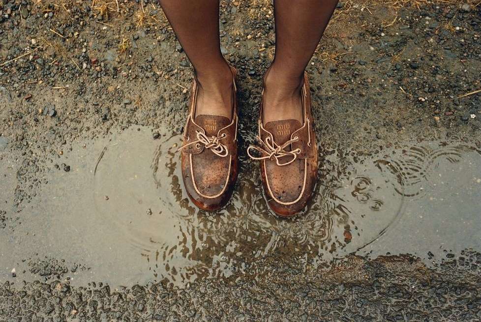 Brown leather boat shoes in a puddle of water, worn with dark tights, showcased in a top-down shot on a gravelly, wet surface.