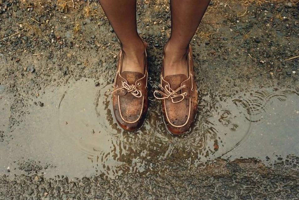 Brown leather boat shoes in a puddle of water, worn with dark tights, showcased in a top-down shot on a gravelly, wet surface.