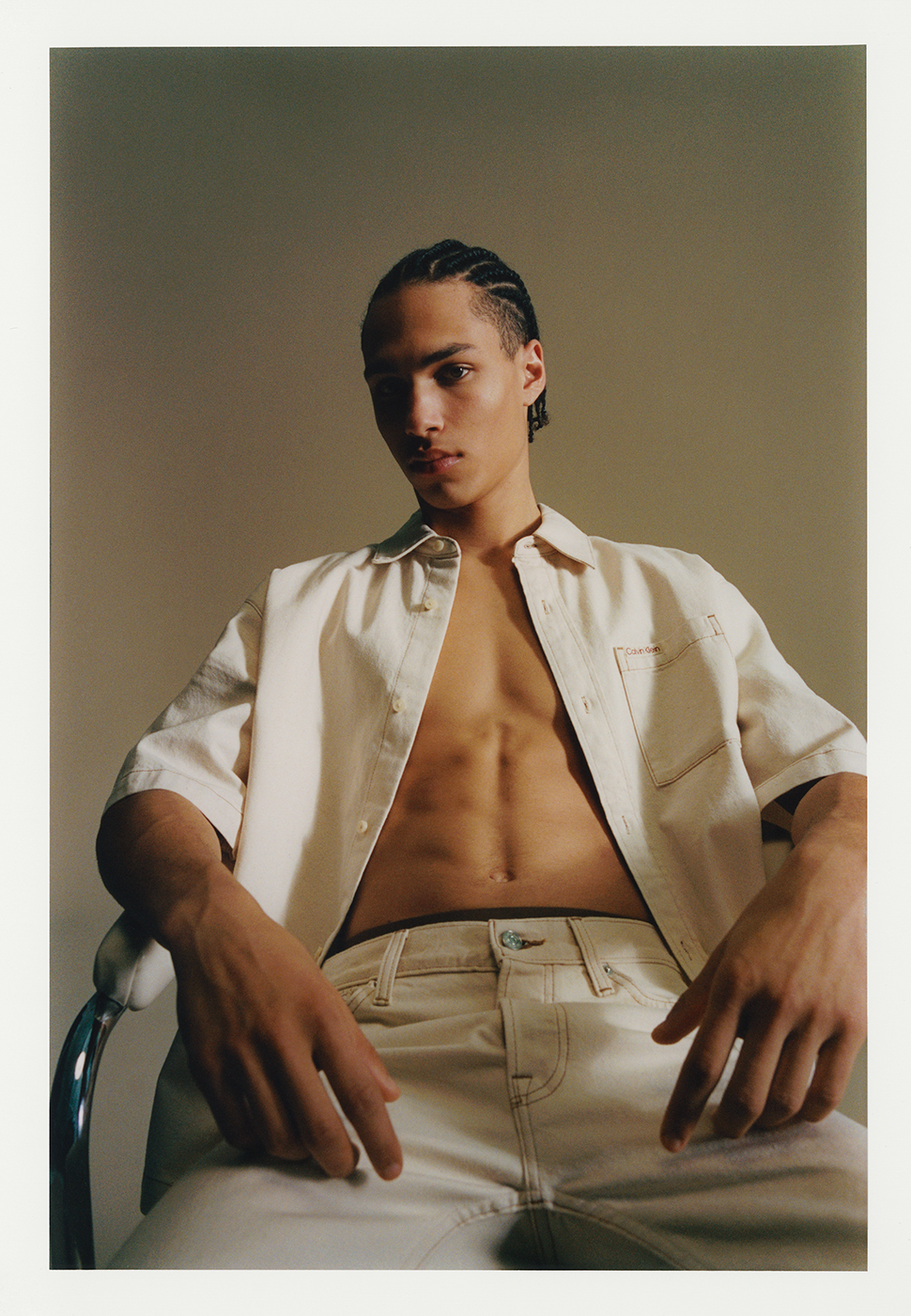Portrait of a young adult male with braided hair sitting on a metal chair, wearing a cream-colored button-down shirt and jeans in studio.