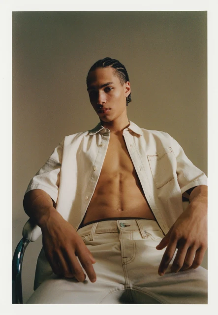 Portrait of a young adult male with braided hair sitting on a metal chair, wearing a cream-colored button-down shirt and jeans in studio.