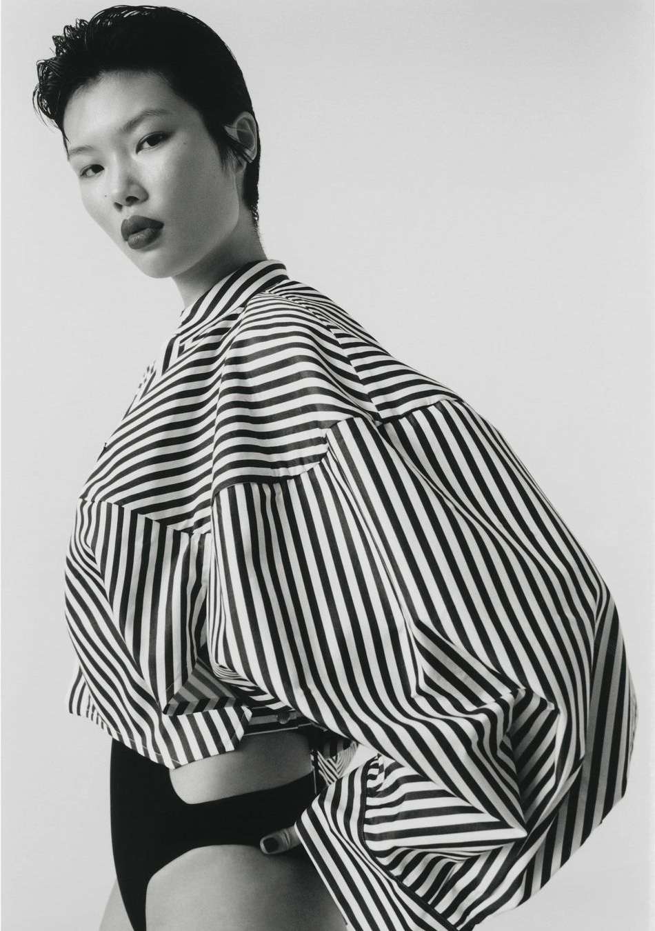 Portrait of an Asian woman in a striped shirt and black bottoms against a white background. Fashion editorial photography.