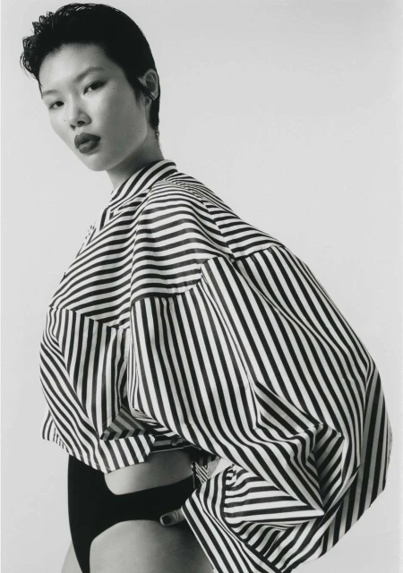 Portrait of an Asian woman in a striped shirt and black bottoms against a white background. Fashion editorial photography.