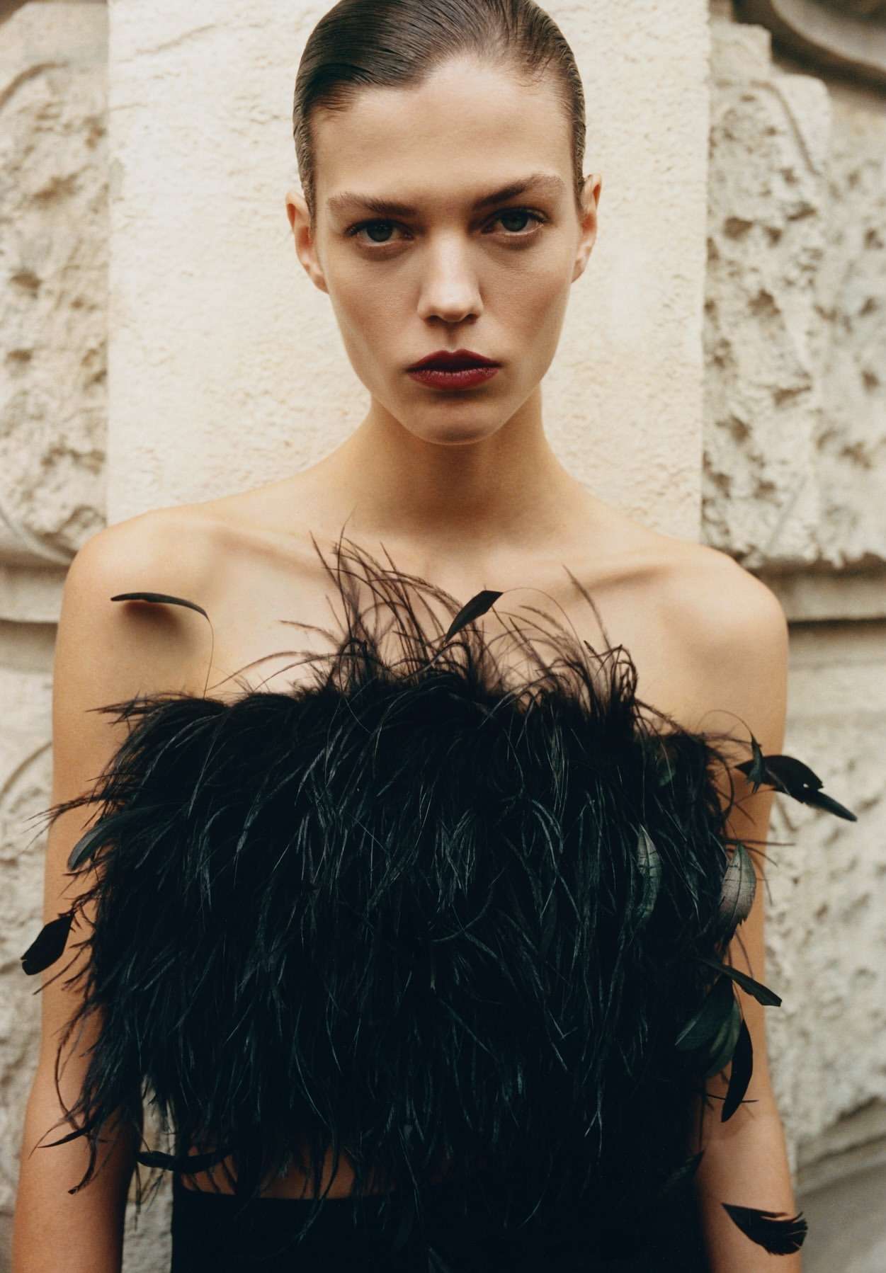 Close-up of a fashion model in a black, feathered top against a neutral background, showcasing beauty and style.