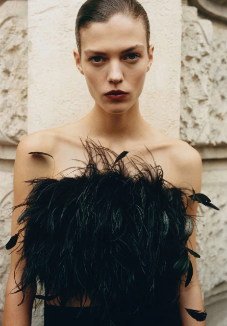 Close-up of a fashion model in a black, feathered top against a neutral background, showcasing beauty and style.