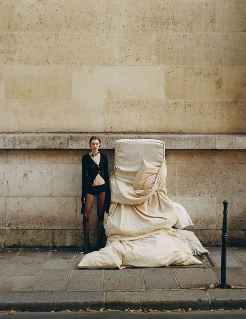 Fashion shot of a slender woman in black lingerie posing next to mattress draped in off-white fabric against a stone wall in urban setting.