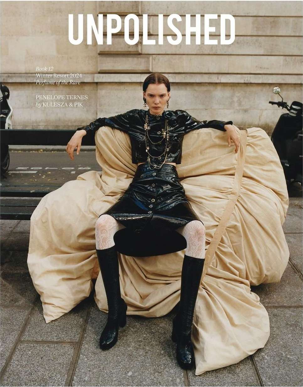 A fashion magazine cover features a model in a black vinyl outfit sitting on a tan fabric set in an urban setting with a scooter.