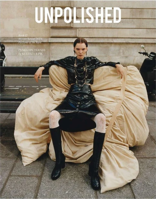 A fashion magazine cover features a model in a black vinyl outfit sitting on a tan fabric set in an urban setting with a scooter.