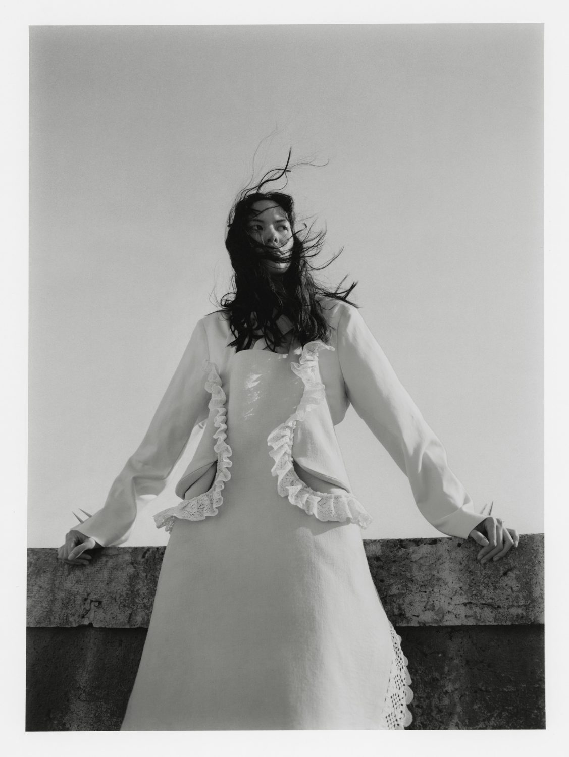 Monochrome portrait of a woman in a ruffled-trim dress and jacket. The breeze catches her hair as she leans on a concrete ledge.