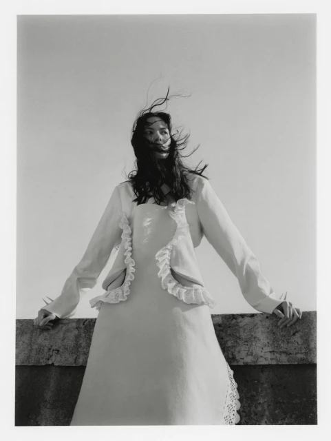 Monochrome portrait of a woman in a ruffled-trim dress and jacket. The breeze catches her hair as she leans on a concrete ledge.