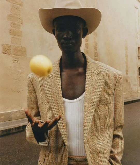 A dark-skinned adult male in a cowboy hat and blazer holds out his hand as a yellow apple floats in front of an outdoor building backdrop.