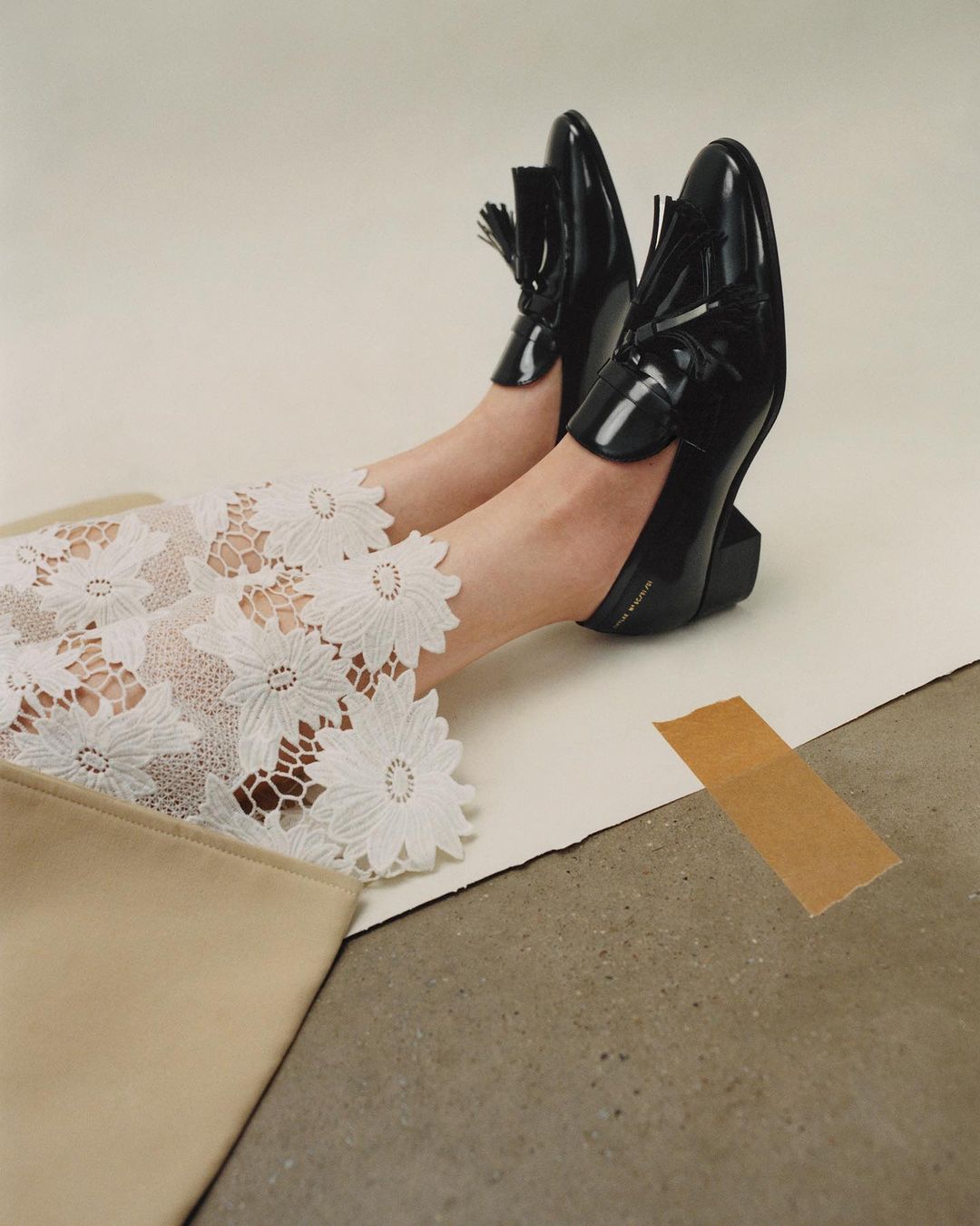 Fashionable black patent leather loafers with tassels on a model's feet, paired with floral lace pants and a beige clutch, studio lit.