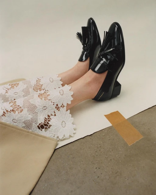 Fashionable black patent leather loafers with tassels on a model's feet, paired with floral lace pants and a beige clutch, studio lit.