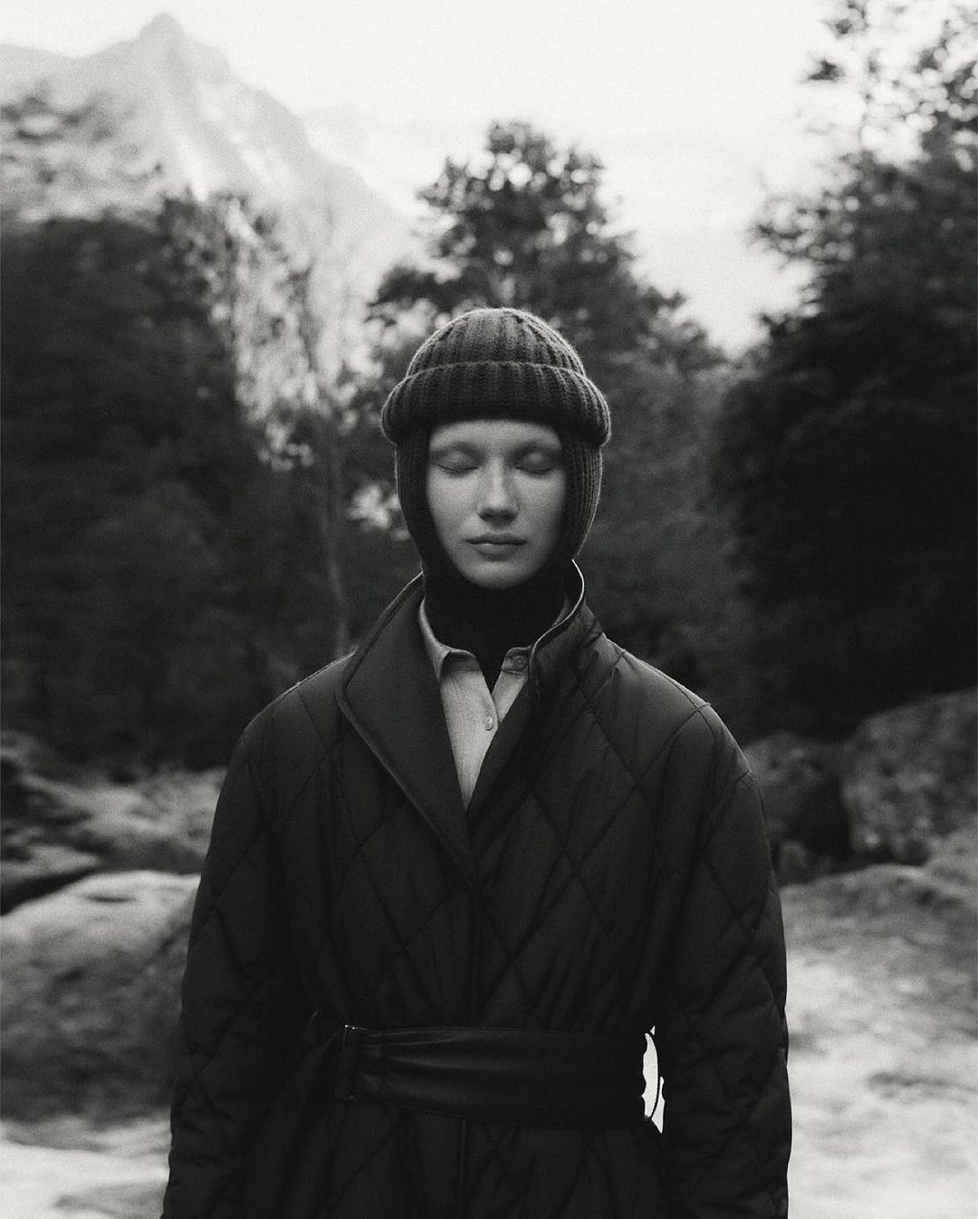 Monochrome portrait of a woman wearing a knit cap and quilted coat outdoors with trees and a mountain in the blurred background.