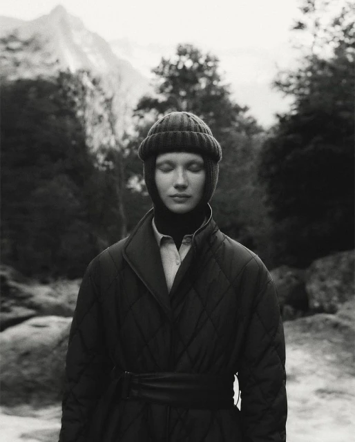 Monochrome portrait of a woman wearing a knit cap and quilted coat outdoors with trees and a mountain in the blurred background.