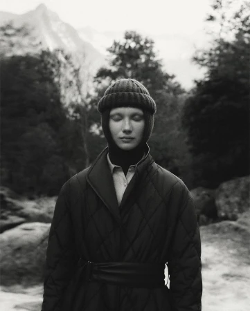 Monochrome portrait of a woman wearing a knit cap and quilted coat outdoors with trees and a mountain in the blurred background.
