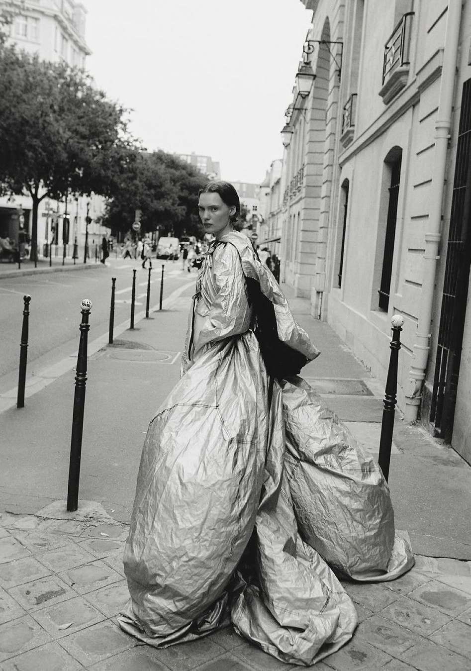 Fashion shot of adult woman in silver couture gown standing on a cobblestone street in front of buildings and street lights in black and white.