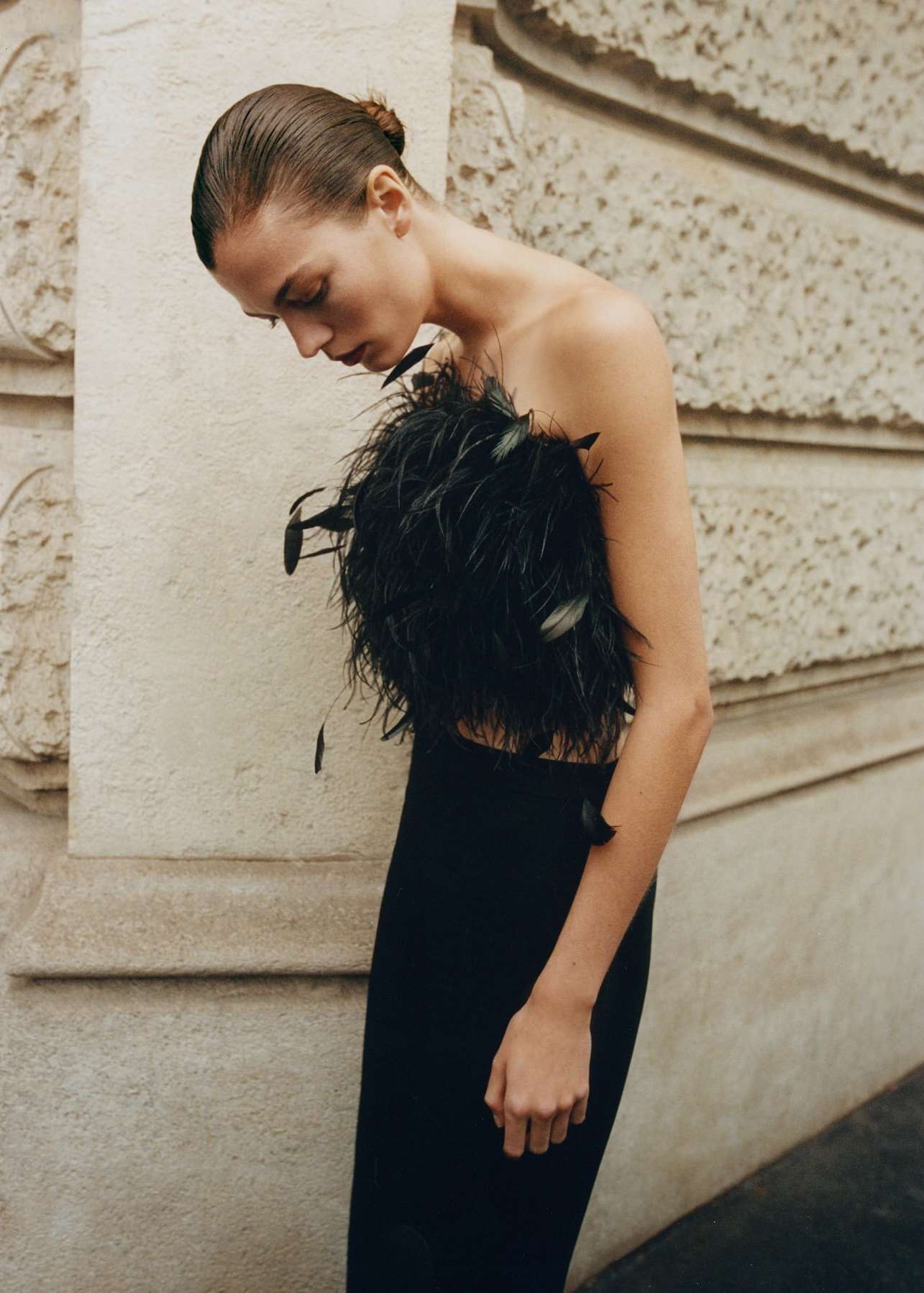 Woman in elegant strapless black feather top and long skirt poses against textured neutral wall in a fashion-style portrait.