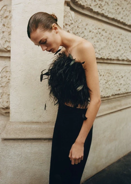 Woman in elegant strapless black feather top and long skirt poses against textured neutral wall in a fashion-style portrait.