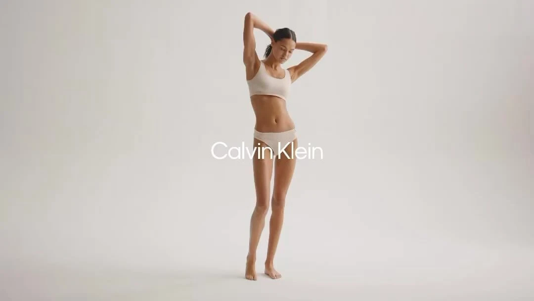 Woman in Calvin Klein underwear poses in a photo studio with hands behind her head in front of a light gray background.