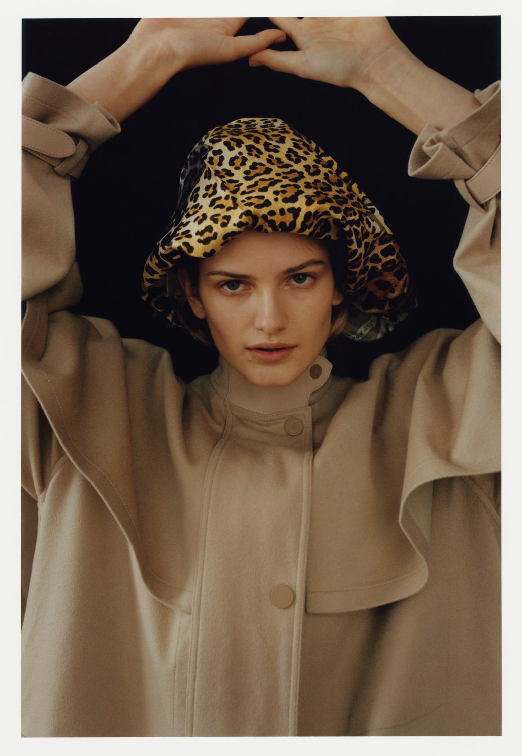 Fashionable woman in neutral trench coat and animal print hat poses against black background in a studio shot with soft, diffused lighting.