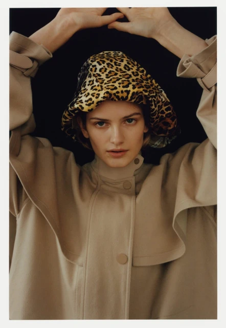 Fashionable woman in neutral trench coat and animal print hat poses against black background in a studio shot with soft, diffused lighting.