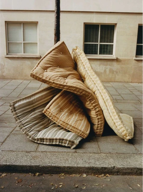 Pile of striped and patterned discarded mattress pads on a gray sidewalk next to a tree and a building with window frames.