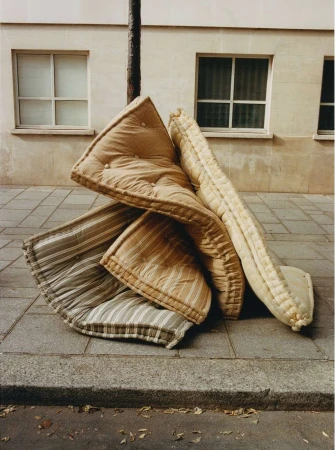 Pile of striped and patterned discarded mattress pads on a gray sidewalk next to a tree and a building with window frames.