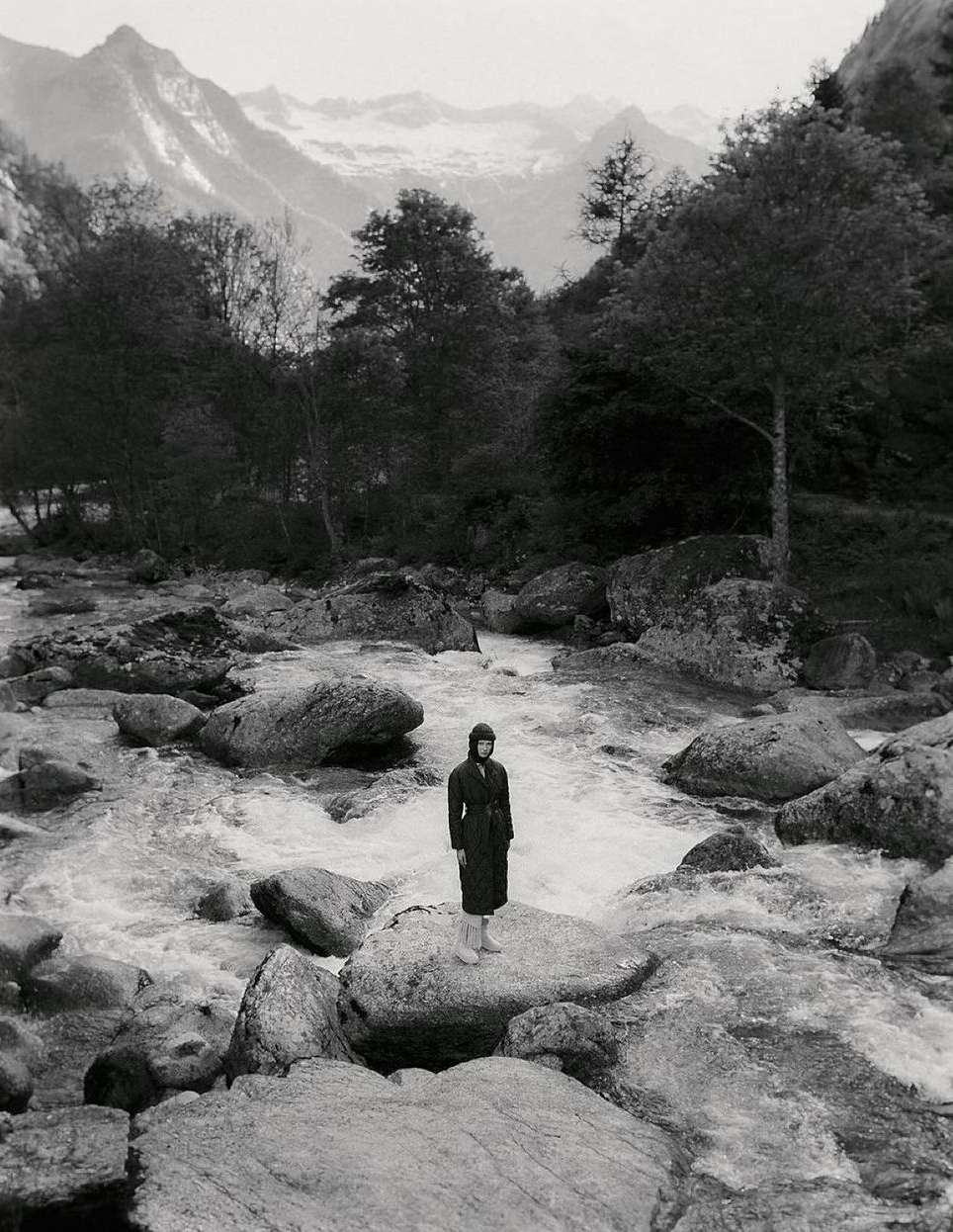 Monochrome landscape featuring a woman standing on a rock in a river, surrounded by lush trees and mountains in the distance, creating a serene nature scene.