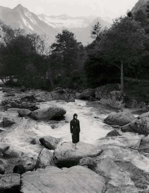Monochrome landscape featuring a woman standing on a rock in a river, surrounded by lush trees and mountains in the distance, creating a serene nature scene.