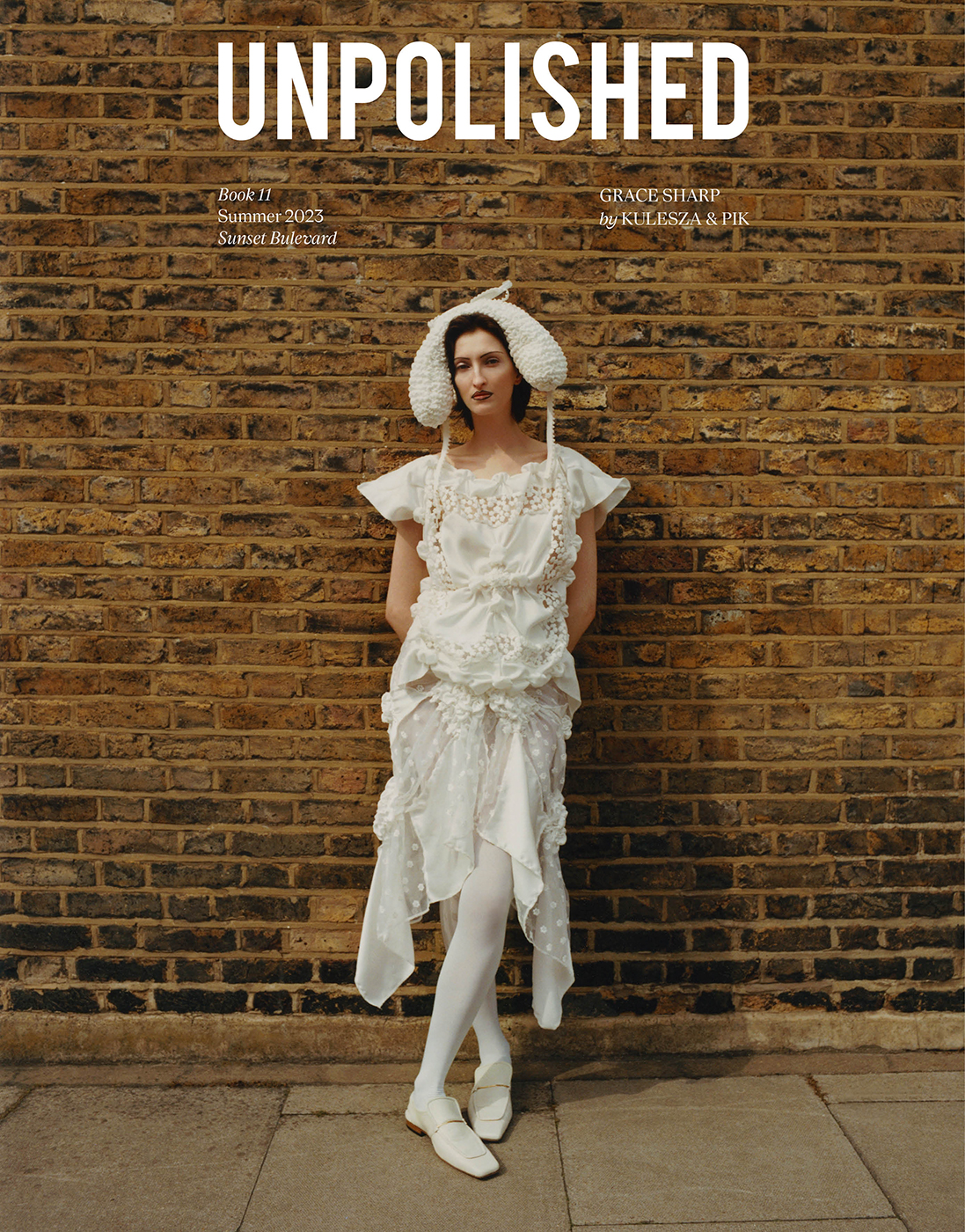 Magazine cover photo of a model in a white dress and decorative hat standing against a brick wall in natural daylight. 