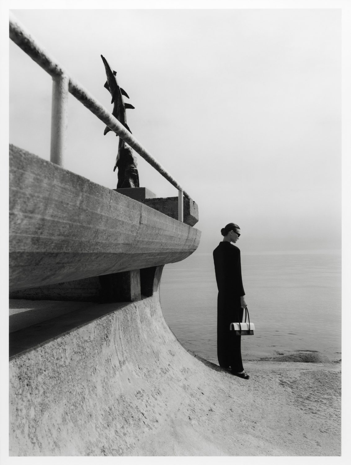 Monochrome fashion shot: Elegant woman in dark suit with purse stands by seaside railing and sculpture. Calm sea and cloudy sky.