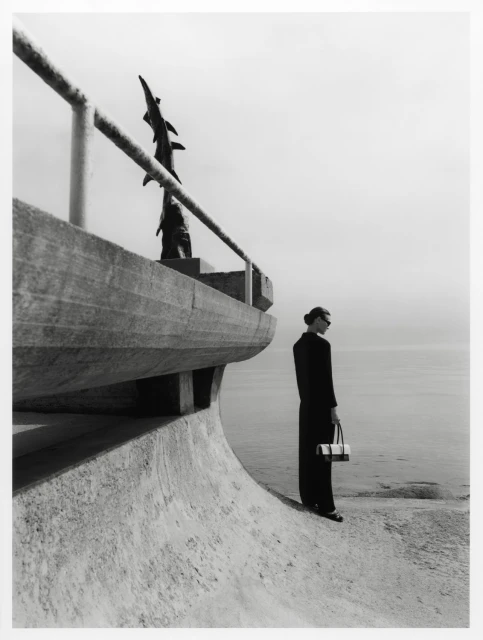 Monochrome fashion shot: Elegant woman in dark suit with purse stands by seaside railing and sculpture. Calm sea and cloudy sky.