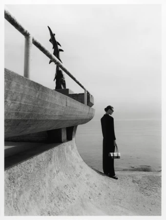 Monochrome fashion shot: Elegant woman in dark suit with purse stands by seaside railing and sculpture. Calm sea and cloudy sky.