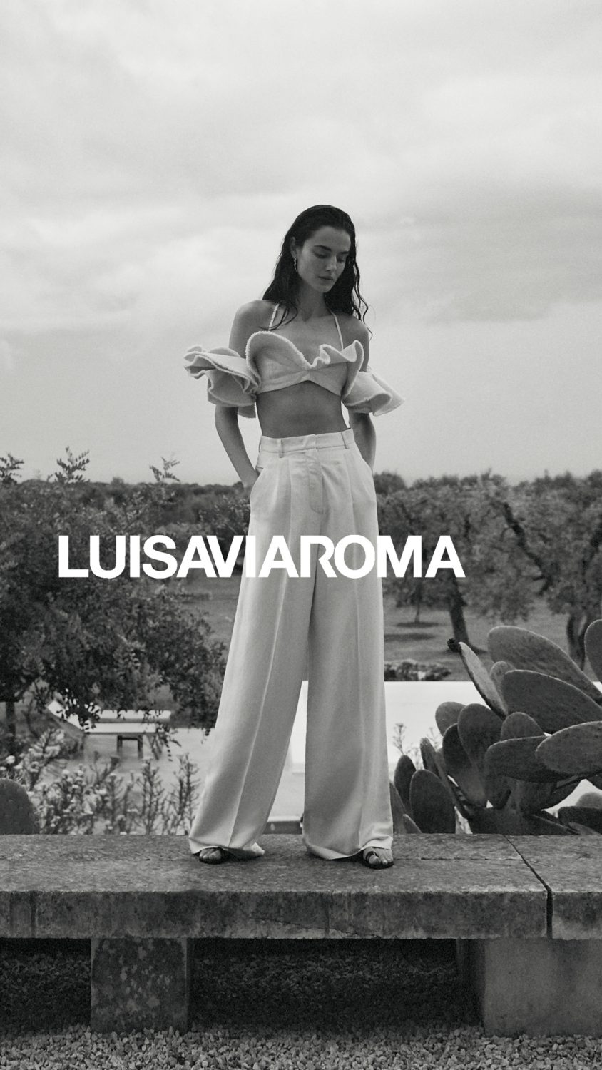 Black and white fashion shot of a young woman in a crop top and linen pants standing on a stone ledge in a rural outdoor setting.